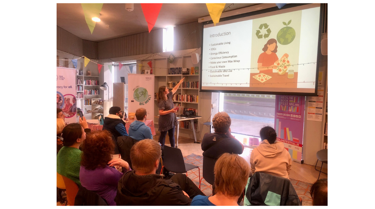 A woman presents a slide about sustainable living to an audience in a decorated library, with banners and posters in the background.